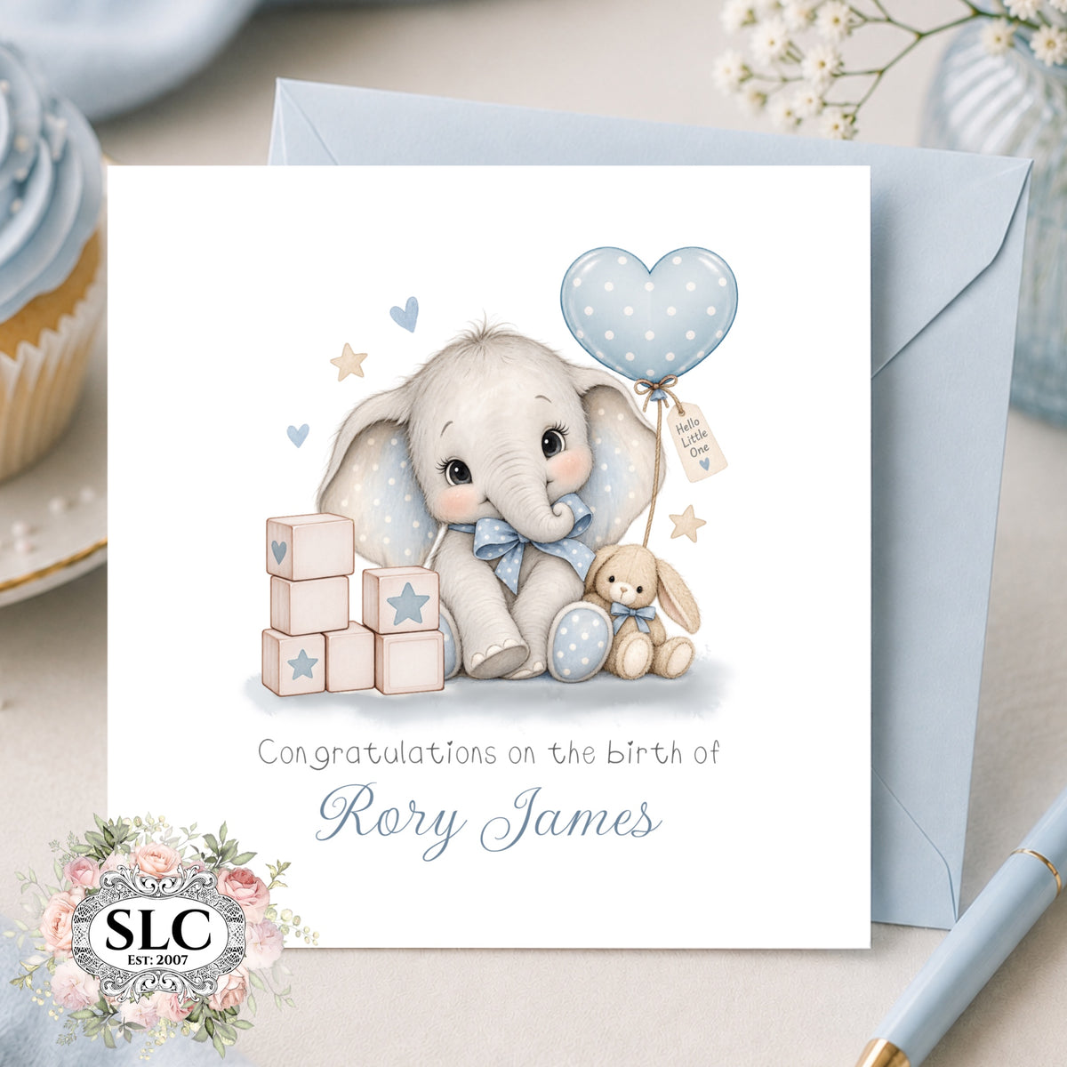 A personalized baby birth announcement card featuring a cute cartoon elephant and bunny with a blue polka-dot balloon, shown with a light blue envelope on a styled tabletop. The card reads 'Congratulations on the birth of Rory James' and includes an 'SLC Est.