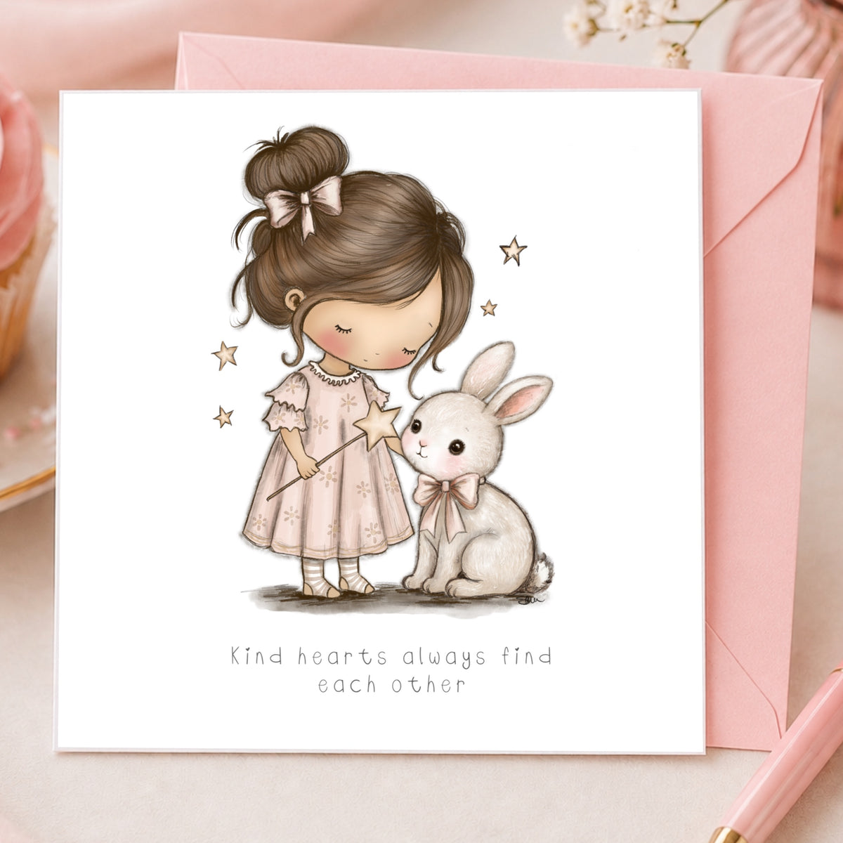 A square greeting card with a white front, featuring an illustration of a young girl in a pink dress holding a star wand next to a white rabbit, with the text 'Kind hearts always find each other' below. The card is shown with a pink envelope on a soft pink surface. collectable cards illustrated by  scruffy little cat 