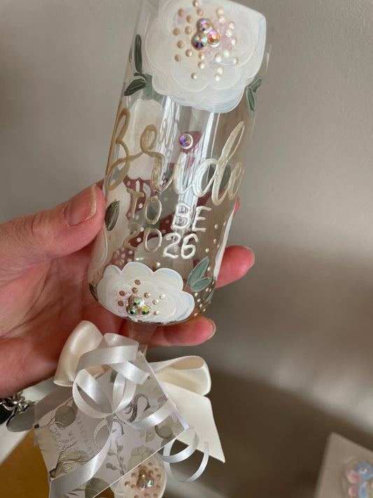 Decorative hand painted champagne flute with bride to be 2026 with floral designs and ribbons held by a hand against a neutral background