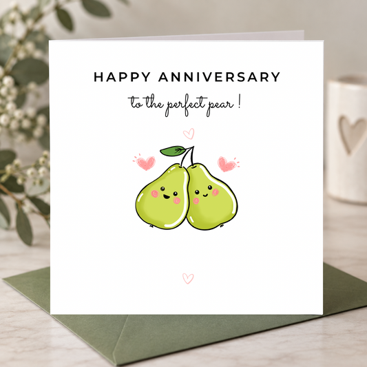A square white anniversary greeting card with the text 'HAPPY ANNIVERSARY to the perfect pear!' and a cute illustration of two smiling green pears with pink hearts, shown standing on a dark green envelope on a light tabletop.