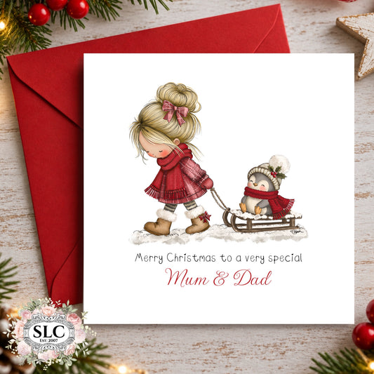 A personalized Christmas greeting card for Mum and Dad, featuring an illustration of a blonde girl in a red dress pulling a penguin in a sled. The card is white with red and black text, shown with a matching red envelope on a rustic wooden background decorated with pine branches and red berries.