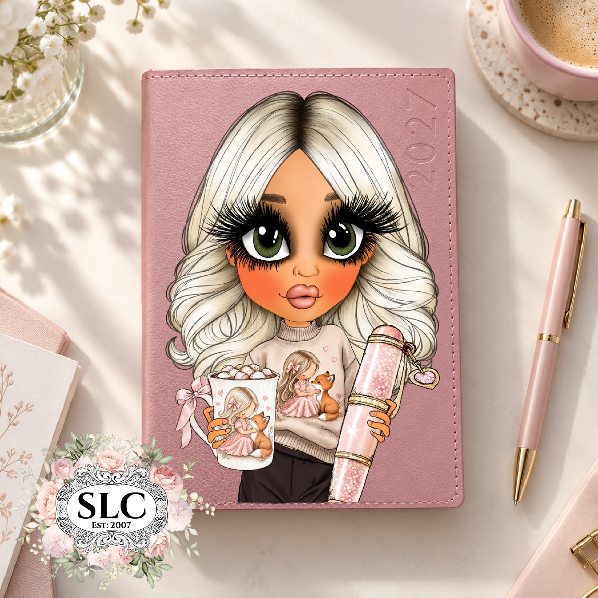 A pink 2027 leather planner or journal cover featuring a stylized cartoon illustration of a blonde woman with large eyes holding a cup and a rolled notebook. The cover is shown on a styled desk flat-lay with a pink pen, coffee cup, and floral accents.
