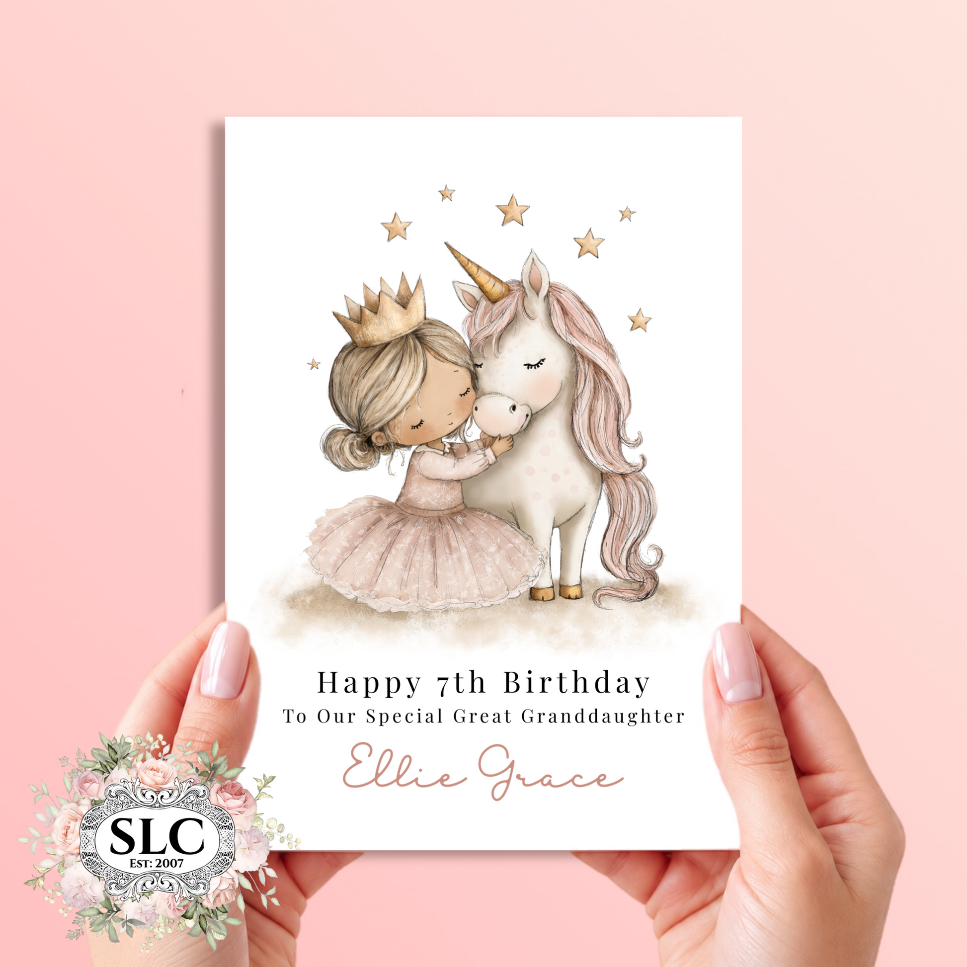 A personalized birthday card held by two hands against a soft pink background. The card features a watercolor illustration of a young girl in a pink dress and gold crown hugging a white unicorn with a gold horn, surrounded by small gold stars.