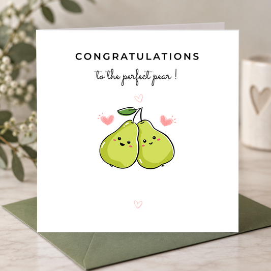 A square white greeting card with the text 'CONGRATULATIONS to the perfect pear!' and a cute illustration of two smiling green pears, shown standing upright on a dark green envelope on a light surface with soft decorative background elements.