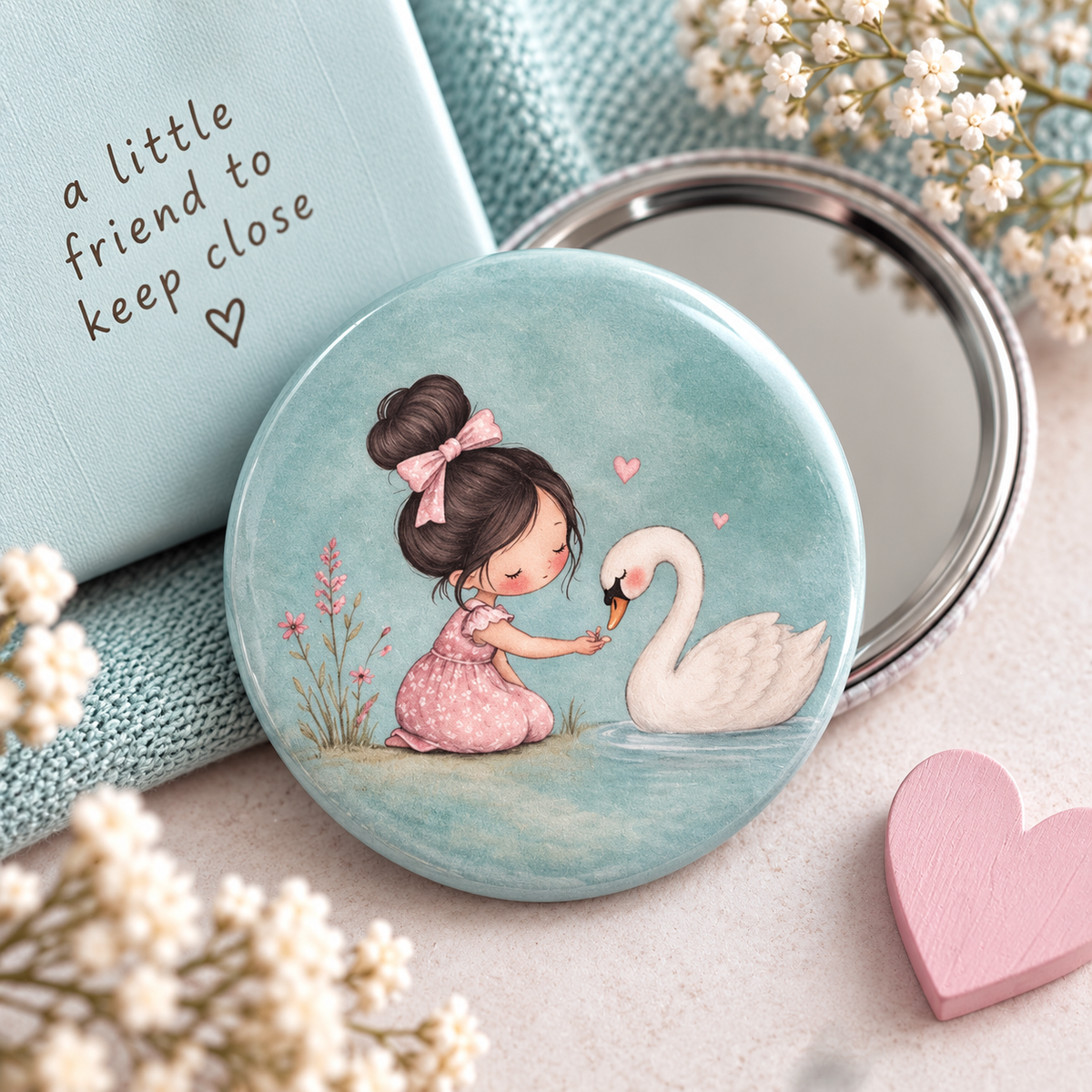 A round compact mirror with a pastel illustration of a girl in a pink dress feeding a white swan a daisy, shown beside a matching light blue box with the text 'a little friend to keep close' and a small pink heart on a soft neutral surface.