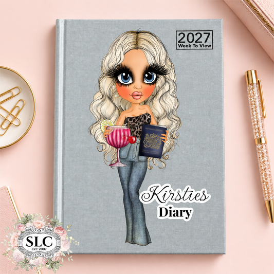 A 2027 Week To View daily diary with a gray cover, featuring a cartoon illustration of a blonde woman holding a pink cocktail and a passport, with the text 'Kirsties Diary' on the front. The diary is styled on a pink background with a gold pen, a paperclip, and a decorative 'SLC Est.