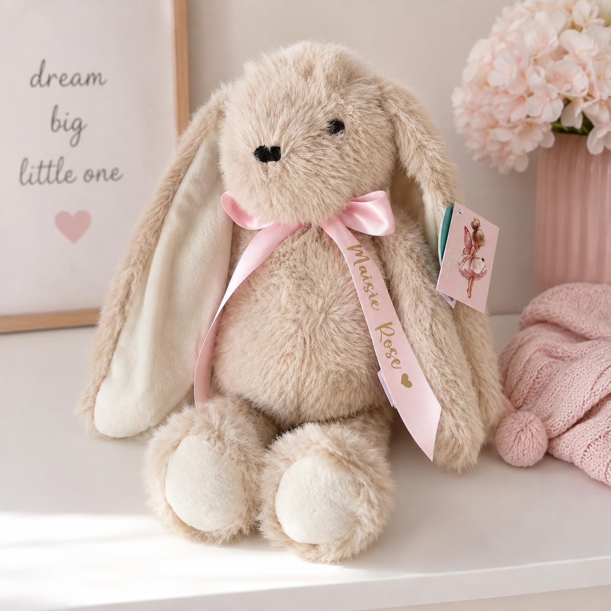 A plush beige bunny stuffed animal with long floppy ears, a pink satin bow, and a hanging  gift tag styled on a white surface with soft nursery decor in the background.