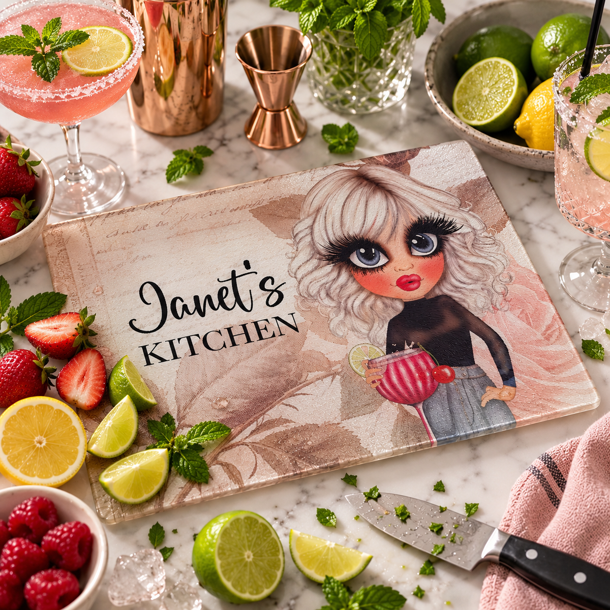 A rectangular decorative cutting board with a cartoon-style blonde woman holding a pink drink and the text 'Janet's KITCHEN,' styled on a marble surface with strawberries, limes, raspberries, mint, copper jigger, glasses, and a knife around it.